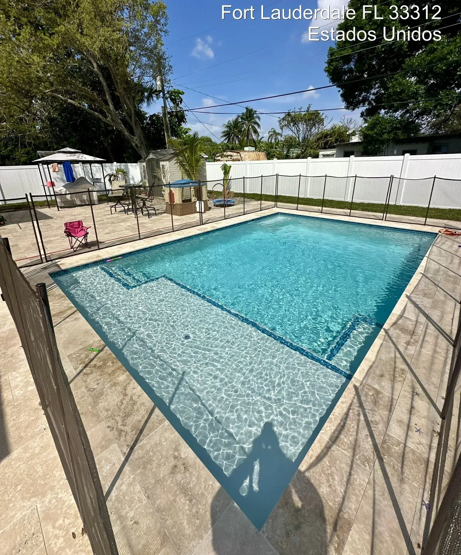 Swimming pool in Fort Lauderdale, Florida, with a fence, steps, and surrounding patio. Blue water under a sunny sky.