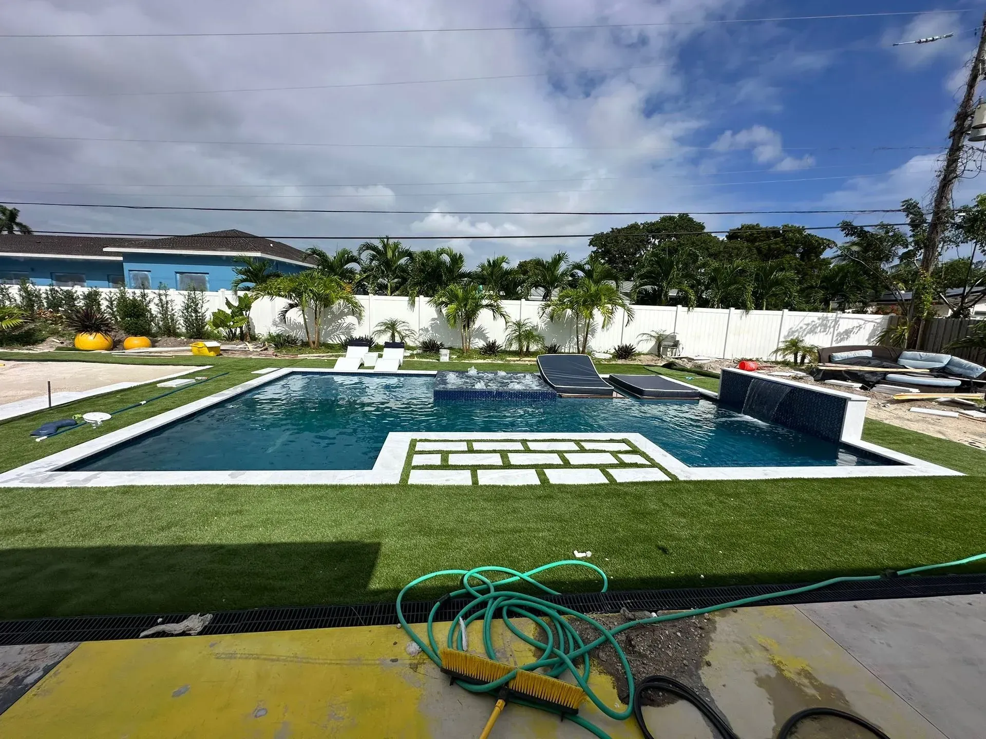Swimming pool with rectangular shape, surrounded by green artificial turf and white tile walkway. Blue sky.