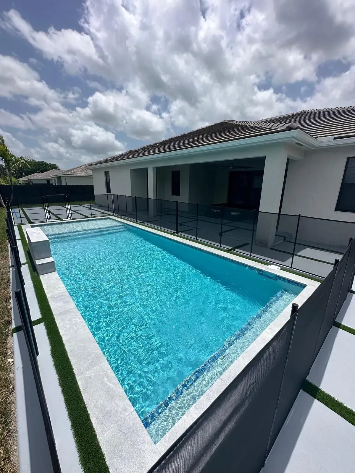 Rectangular blue pool with white trim next to a house with a tiled roof under a cloudy sky.