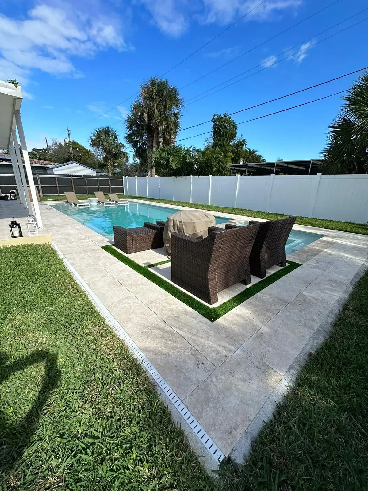 Backyard with pool, patio, and lounge seating; green lawn, white fence, blue sky.