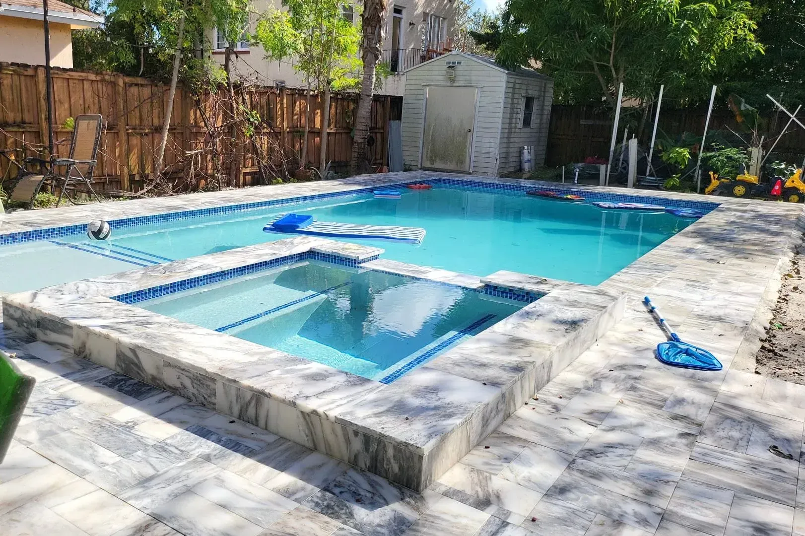 Swimming pool with spa, light blue water, surrounded by gray stone, wooden fence backdrop.