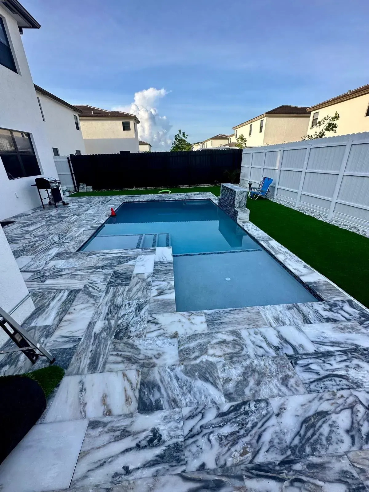 Swimming pool with marble patio, adjacent to a house, with green turf strip along white fence.