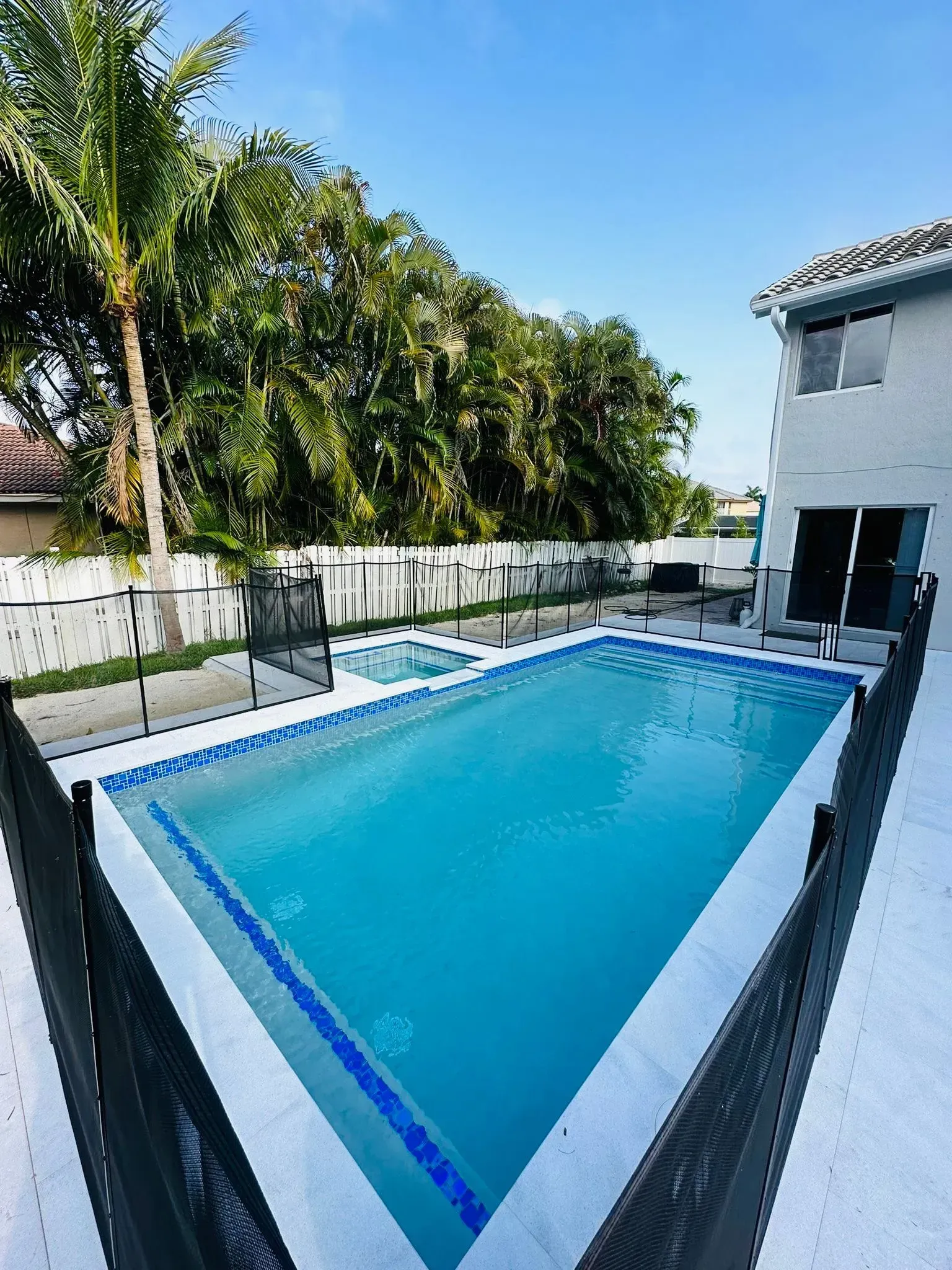 Backyard pool with spa, enclosed by black fence, near a two-story building and lush trees, on a sunny day.
