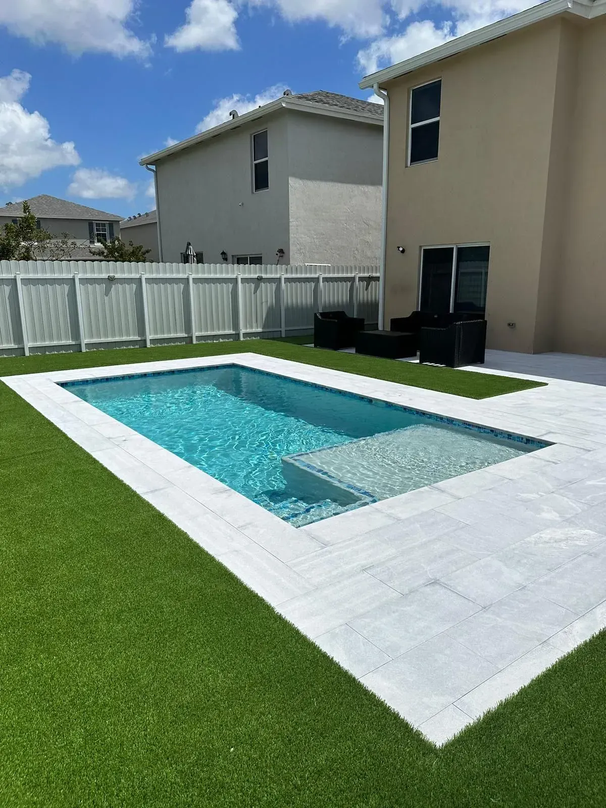 Rectangular swimming pool with white tile surround, green lawn, and beige houses under blue sky.