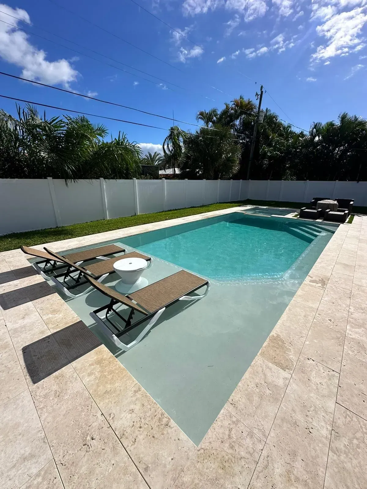 Pool with lounge chairs, blue water, beige patio, and a white fence under a partly cloudy sky.