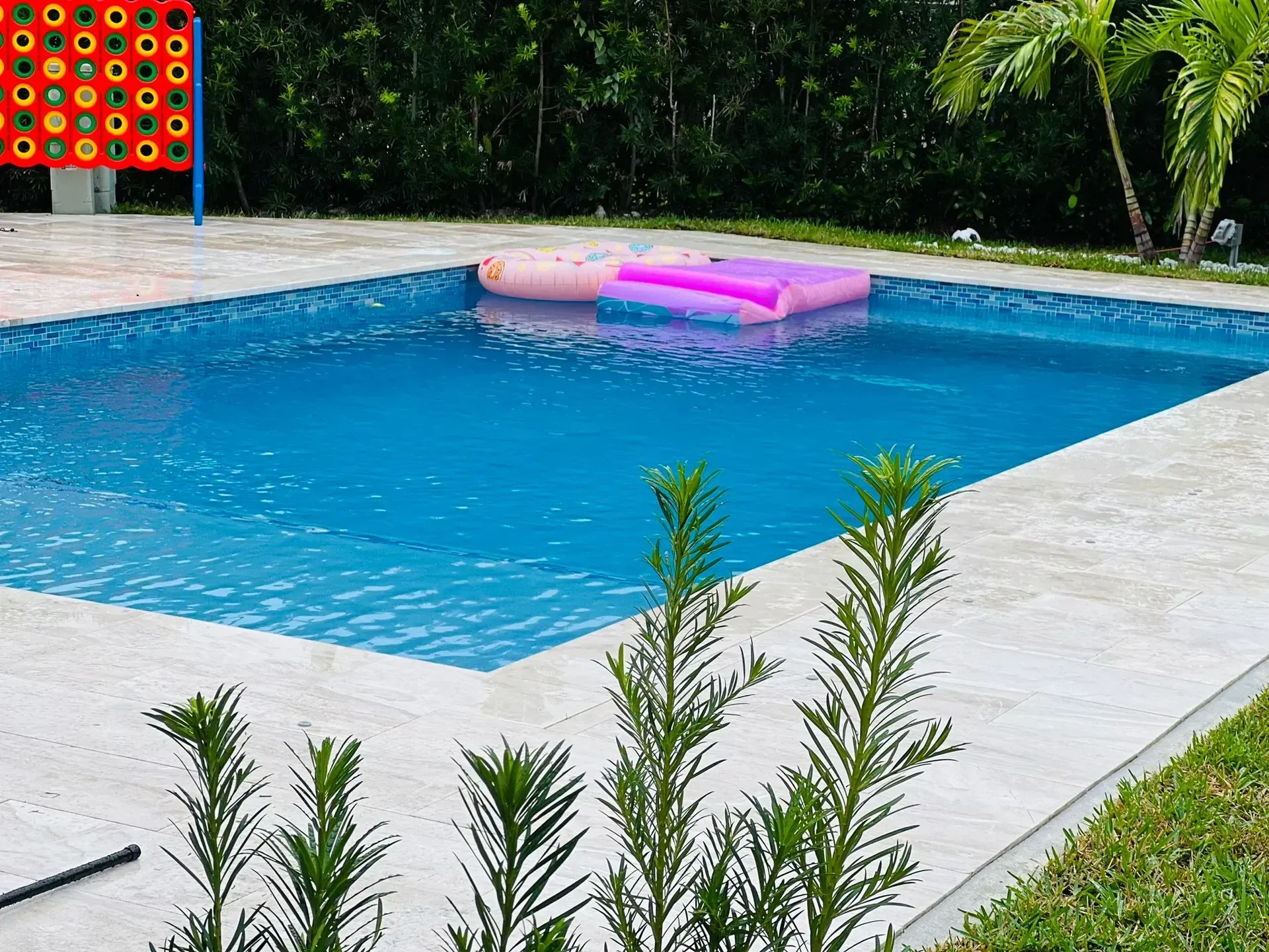 Swimming pool with blue water, white deck, and floaties. A Connect Four game sits in the background.
