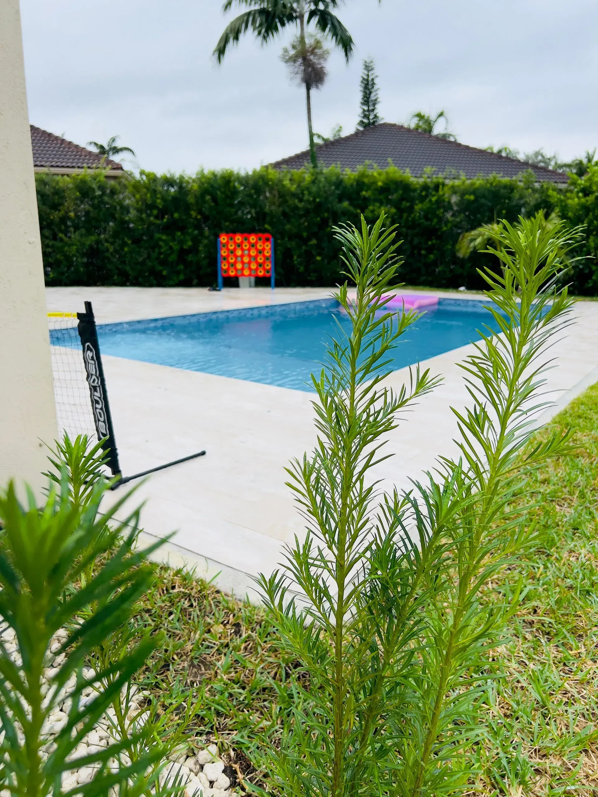 A pool with clear blue water and white concrete is surrounded by grass and greenery. A game is set up near the pool.