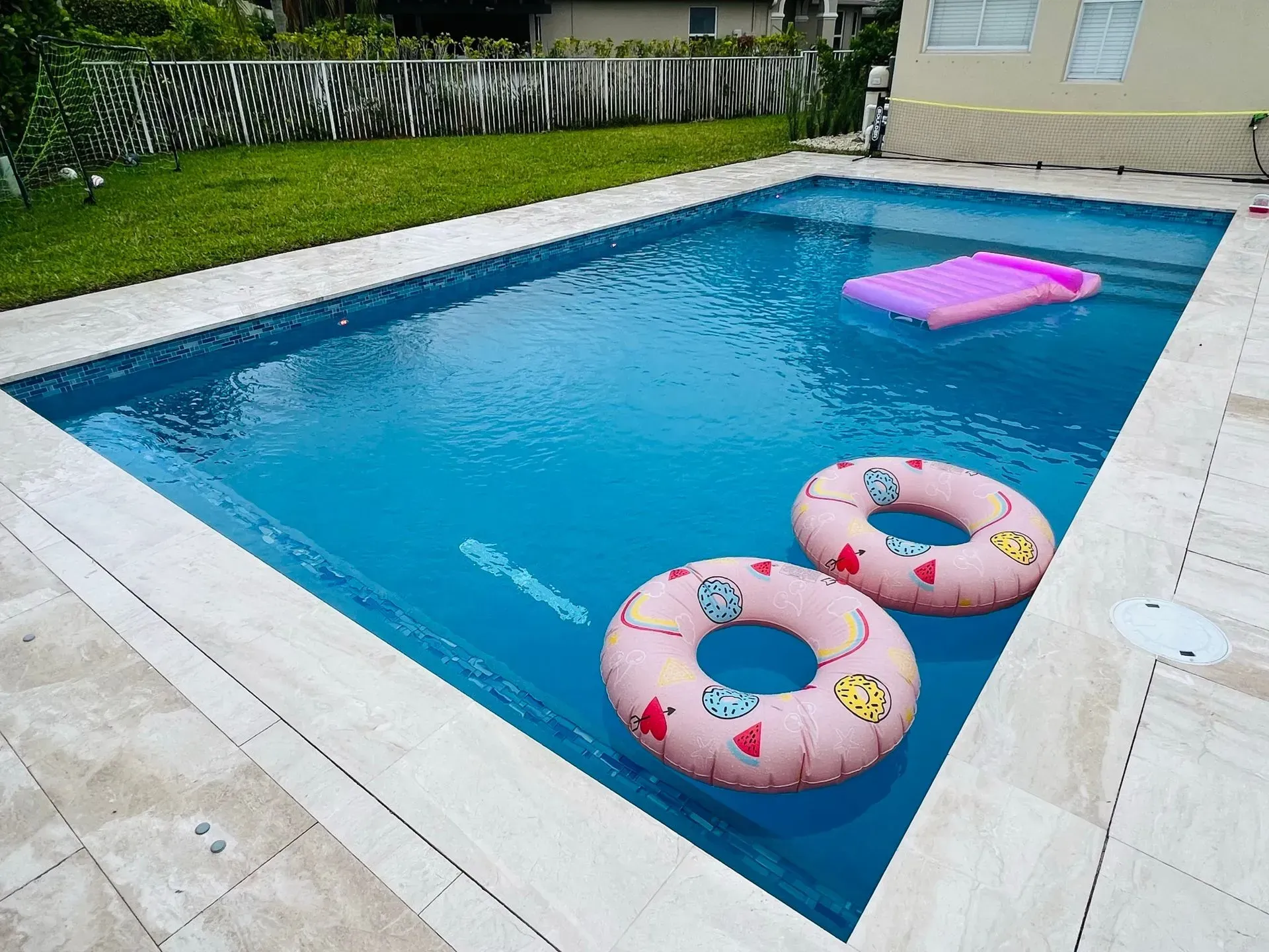 Swimming pool with clear blue water and pool floats. The pool is bordered by light-colored stone. Green lawn in the background.