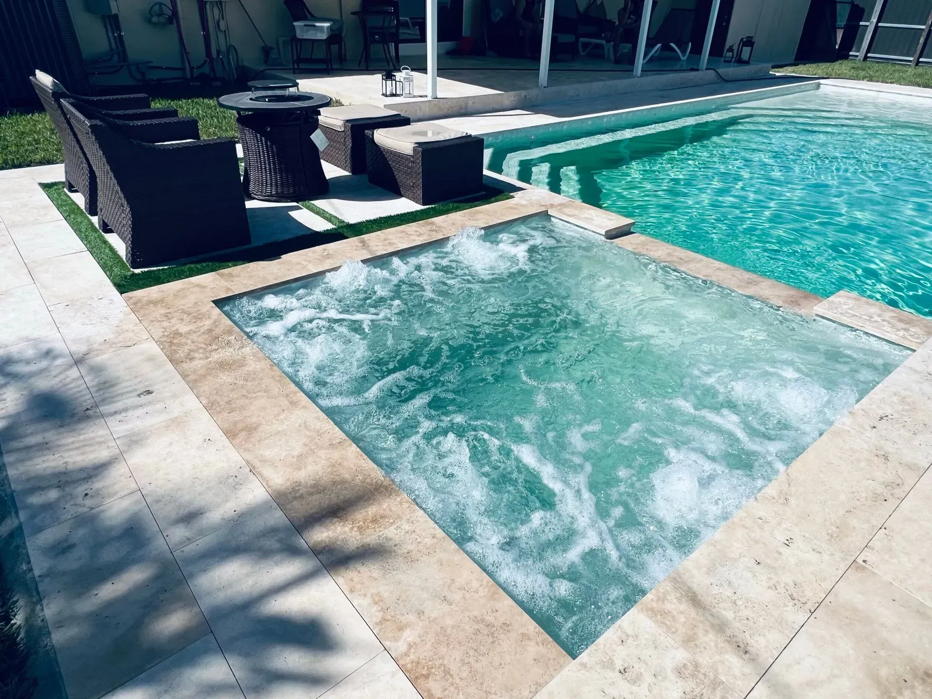Jacuzzi next to a pool with steps, surrounded by light stone. Wicker chairs sit nearby.