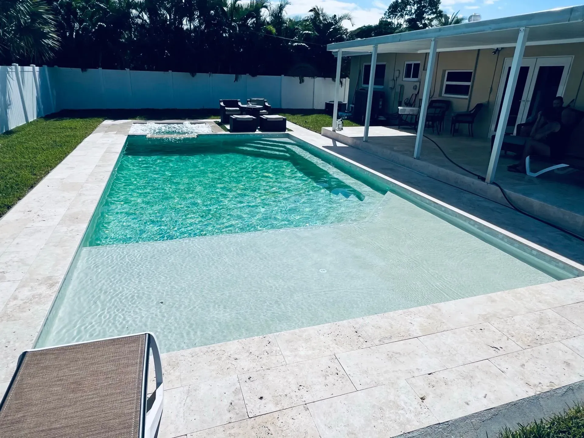 Rectangular turquoise pool with white stone coping, steps, and a shallow sun shelf. Patio with seating nearby.