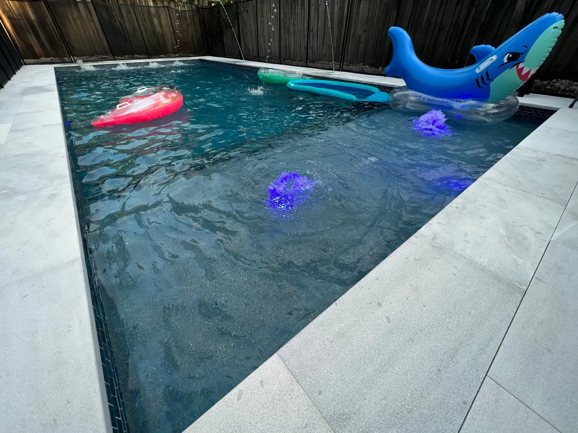 A rectangular swimming pool with floats and lights.  Blue water, white tile, and a wooden fence are visible.