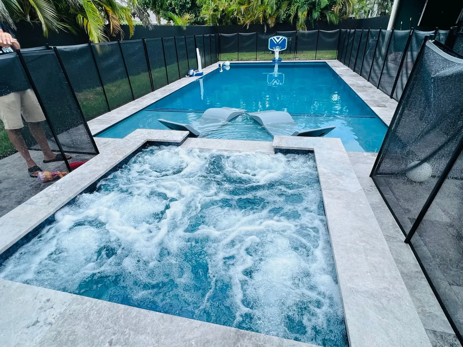 Rectangular pool with attached hot tub, water jets active. Person standing at pool edge. Black fence surrounds.