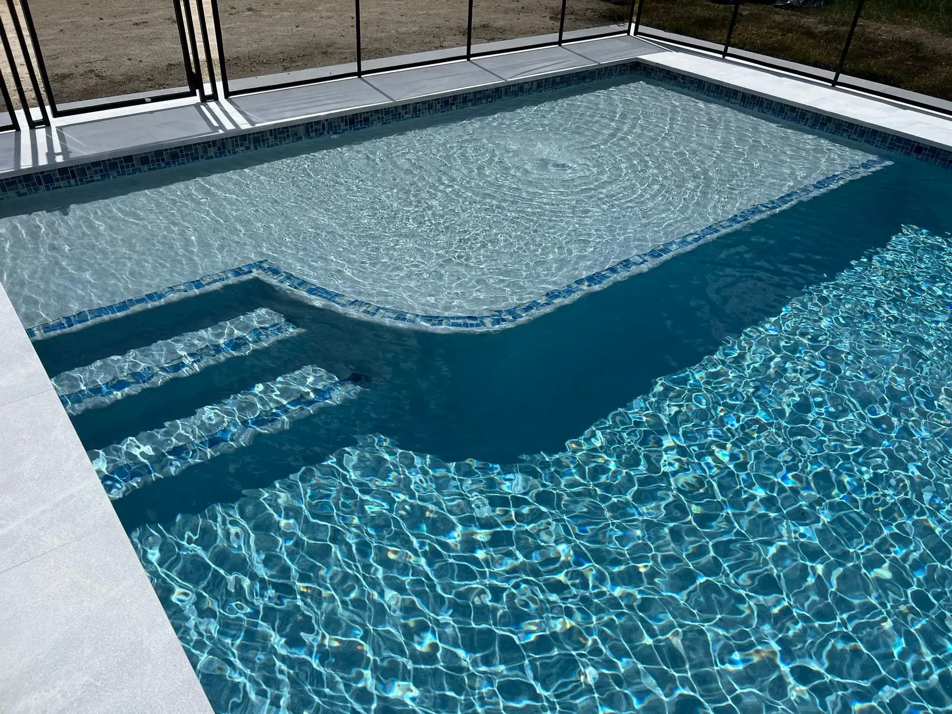 Pool with steps and a shallow area, surrounded by a light-colored patio and black fencing.
