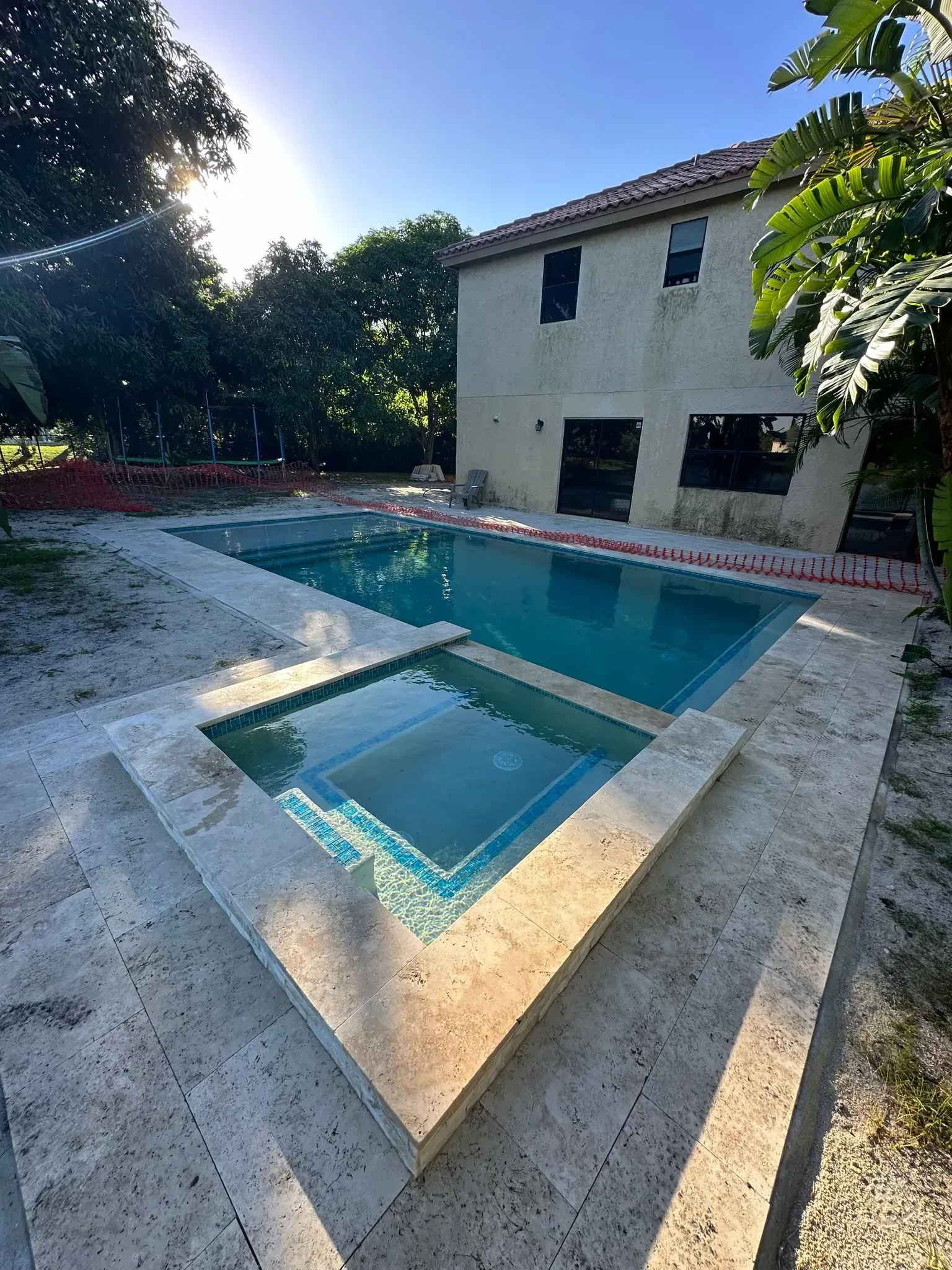 A rectangular swimming pool and a square spa with beige stone surrounding them. A house is in the background.