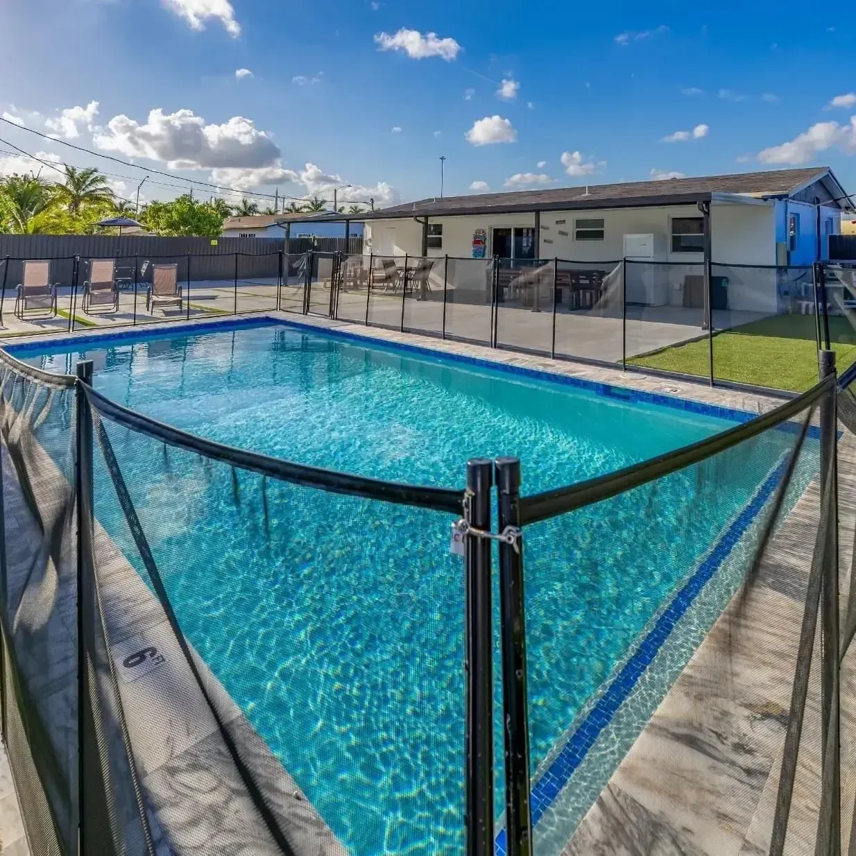 Backyard pool surrounded by a black mesh fence, near a house with a blue sky.