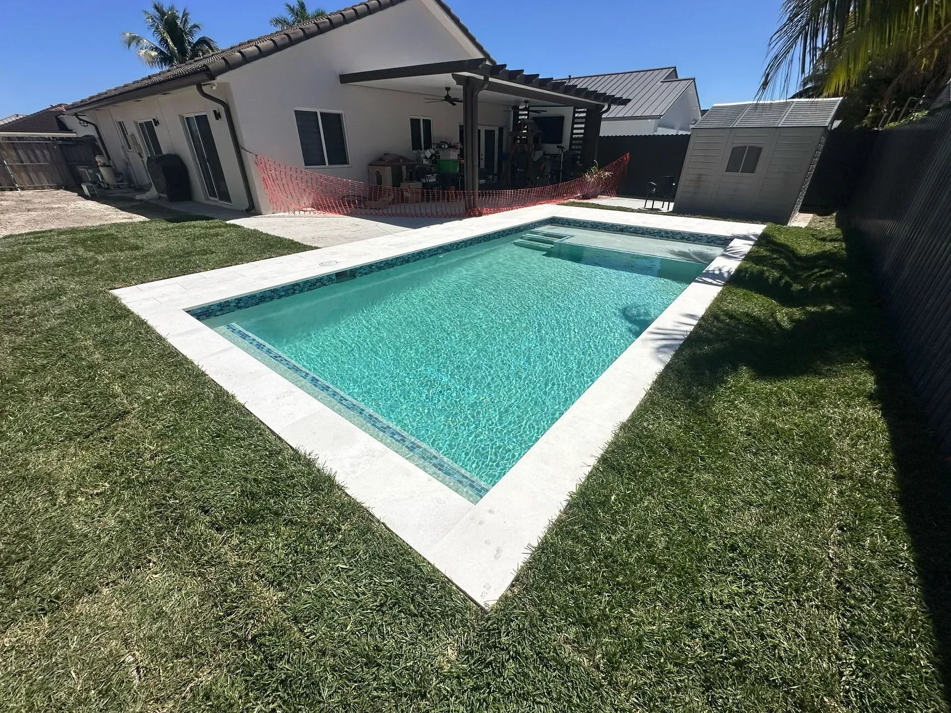 Backyard with a rectangular swimming pool, bordered by white concrete, surrounded by green grass. A house is in the background.
