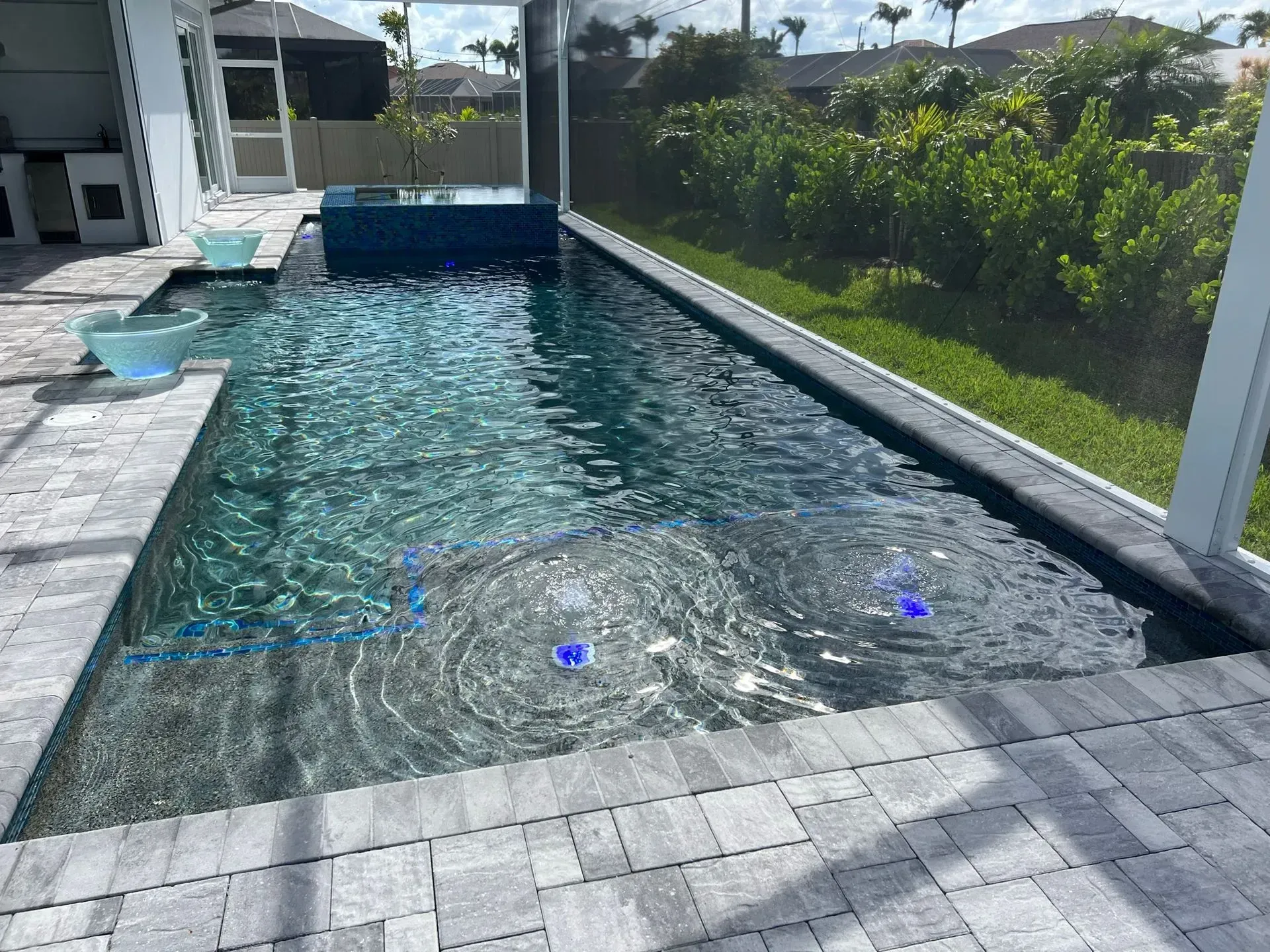 Rectangular outdoor pool with blue-green water surrounded by gray pavers. Two water features are active.