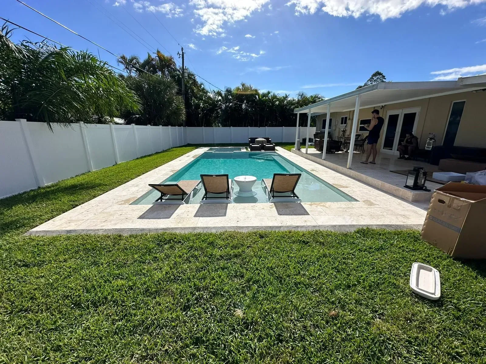 Backyard pool with lounge chairs, white table, and house with covered patio. Blue sky and green grass.