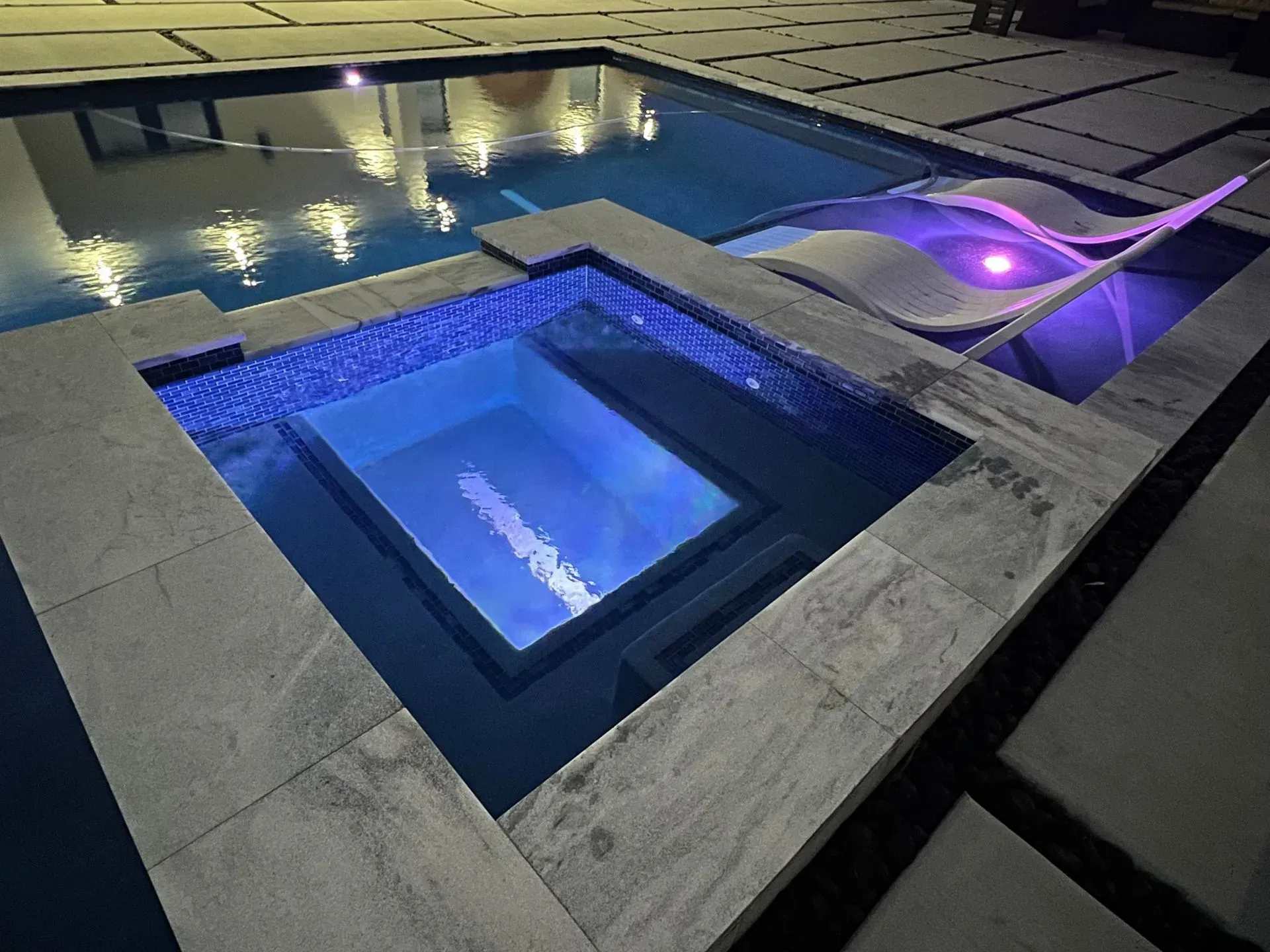A modern pool area with a square hot tub, pool, and lounge area, all illuminated in blue and purple.
