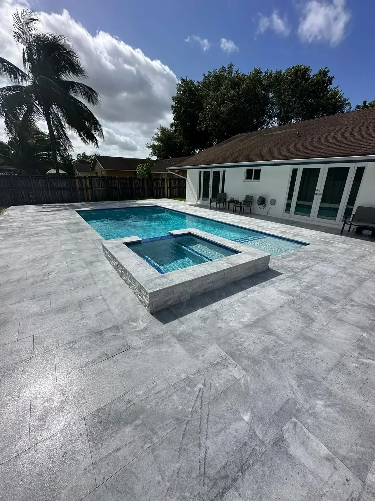 Swimming pool with built-in spa. Gray stone patio, blue water. White house, green trees, sunny sky.