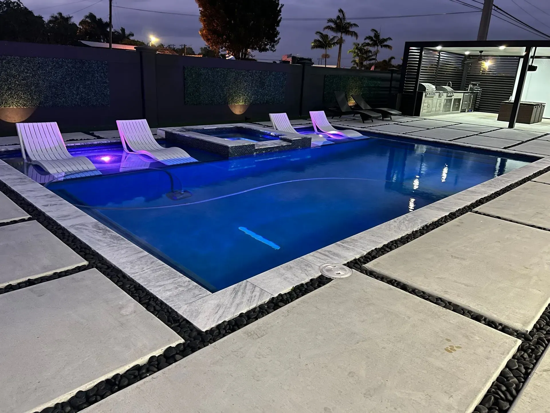 Nighttime view of a rectangular pool with submerged lights, lounge chairs, and a small hot tub. Concrete patio.