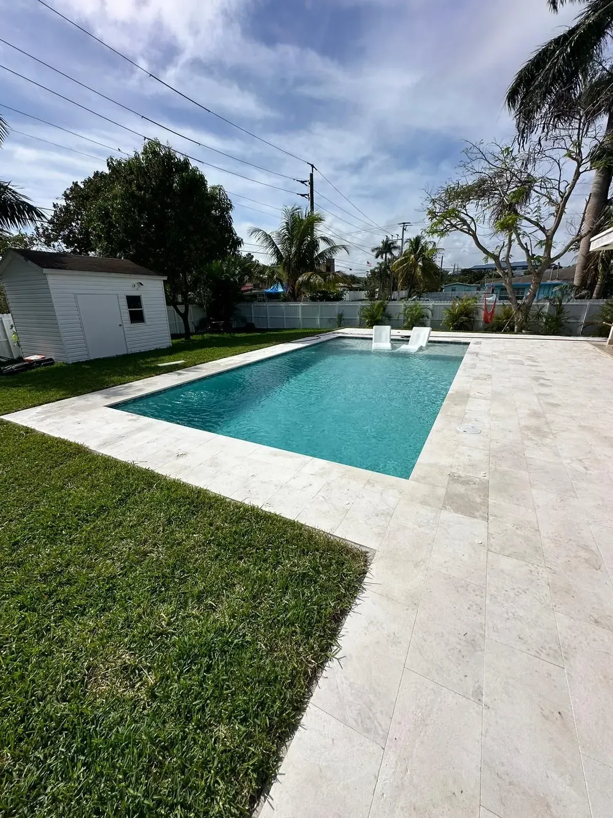 Backyard pool with white stone patio, green grass, and white shed under a sunny sky.