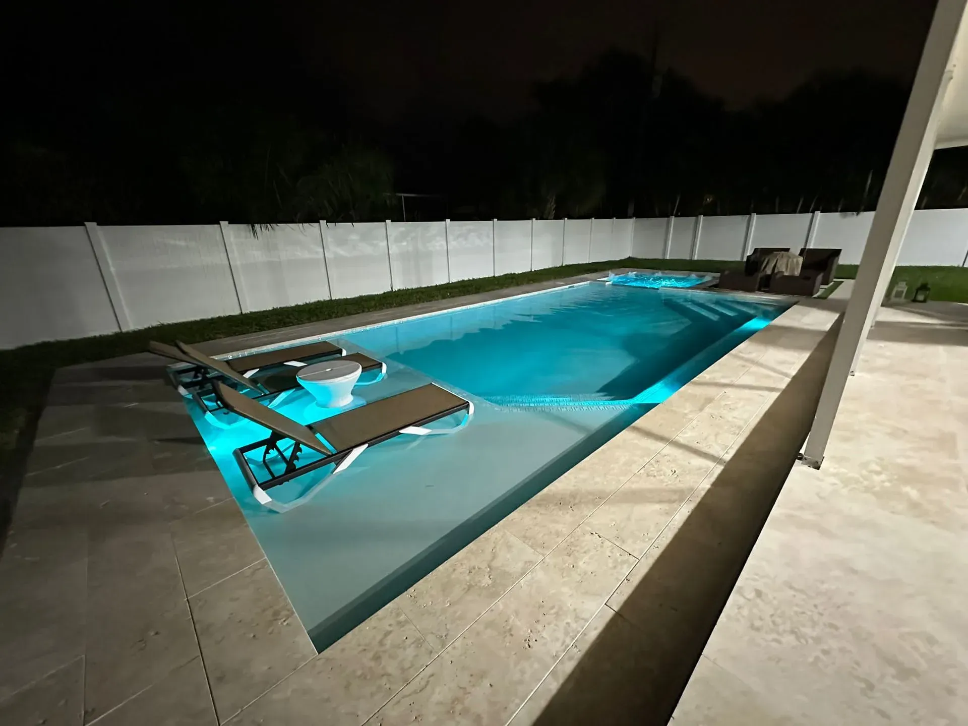 Nighttime view of a rectangular pool illuminated with blue light, lounge chairs, and a white fence.
