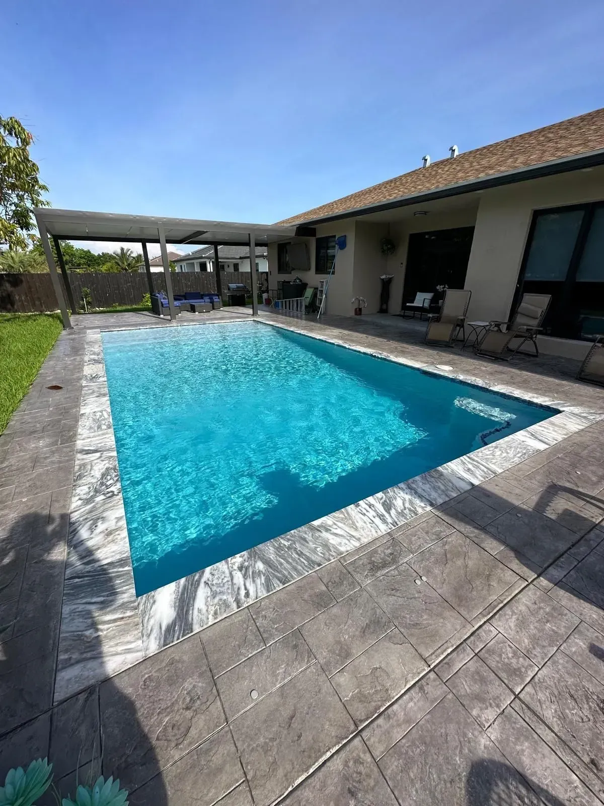 Rectangular swimming pool in a backyard with a covered patio. Blue water, gray concrete, and a clear sky.