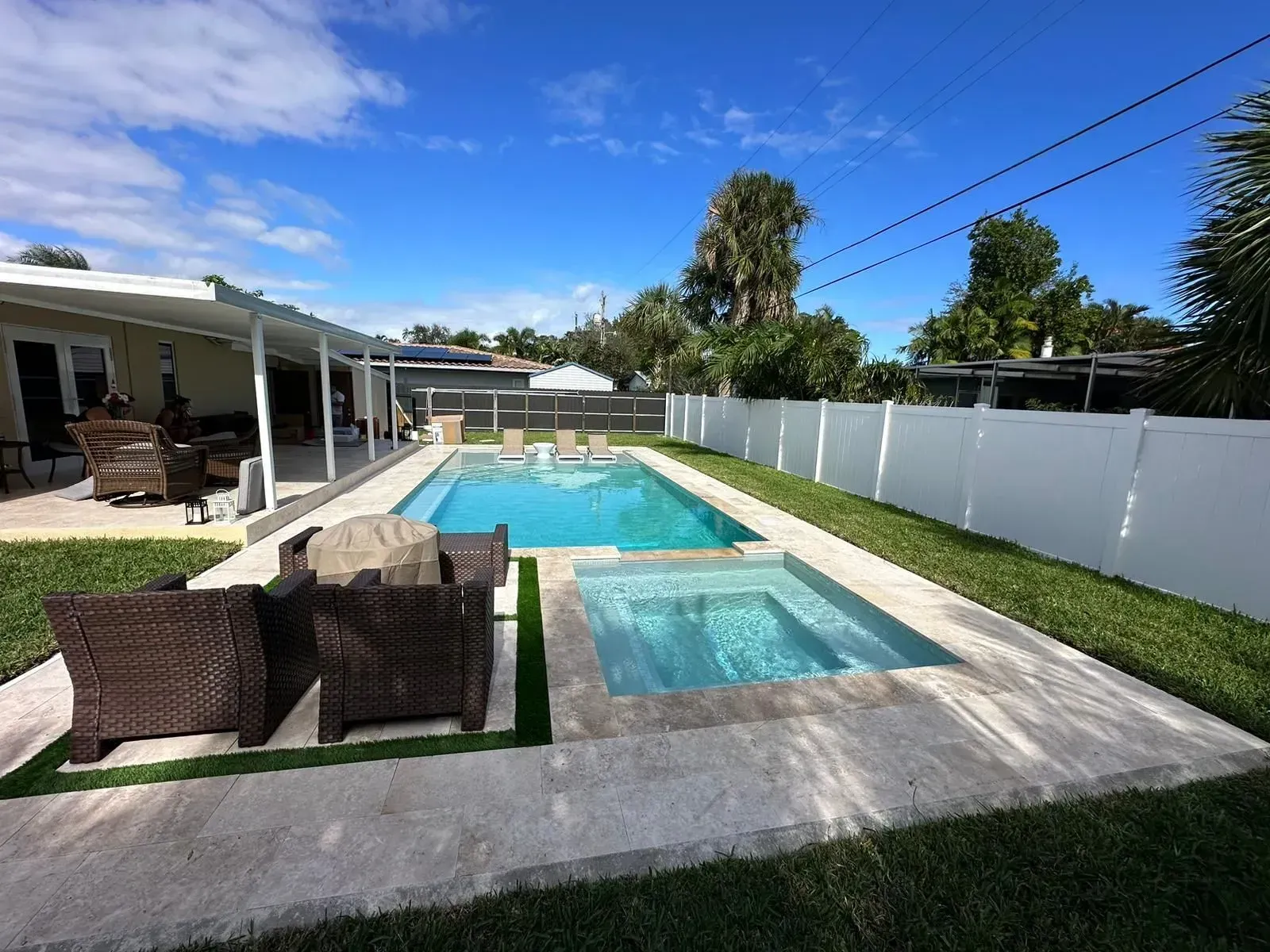 Backyard pool with spa, lounge furniture, patio, and white fence under a blue sky.