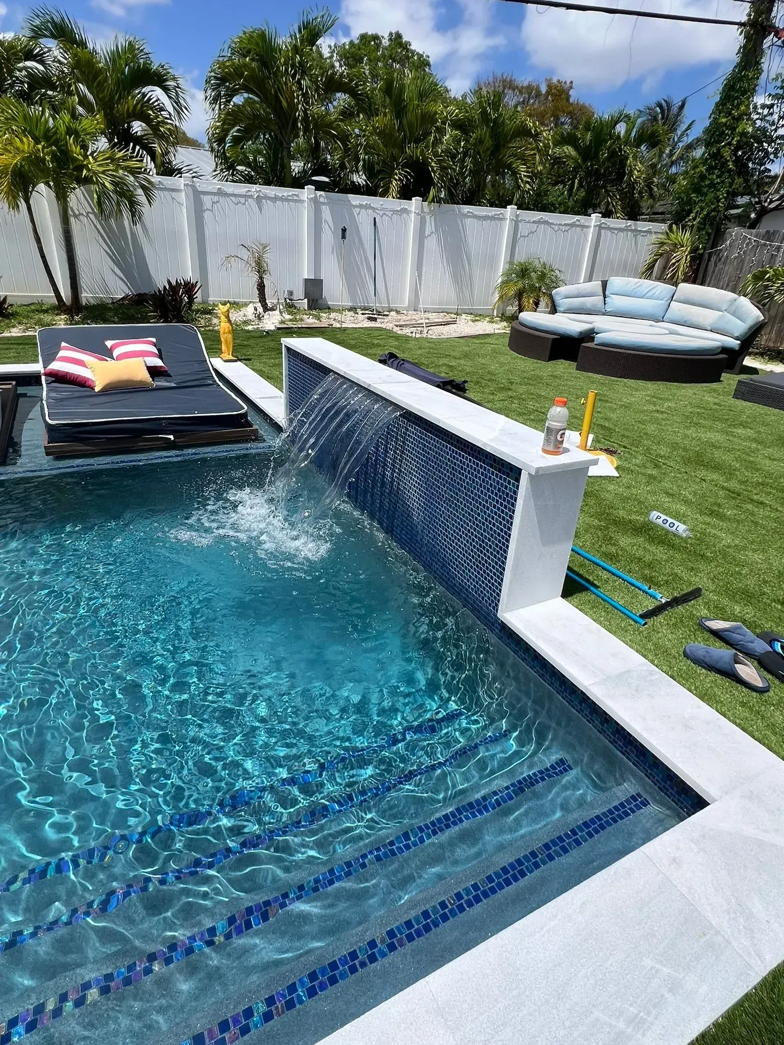 Pool with waterfall feature and blue tiled wall. Lounge chairs and outdoor seating in the background. Green lawn.