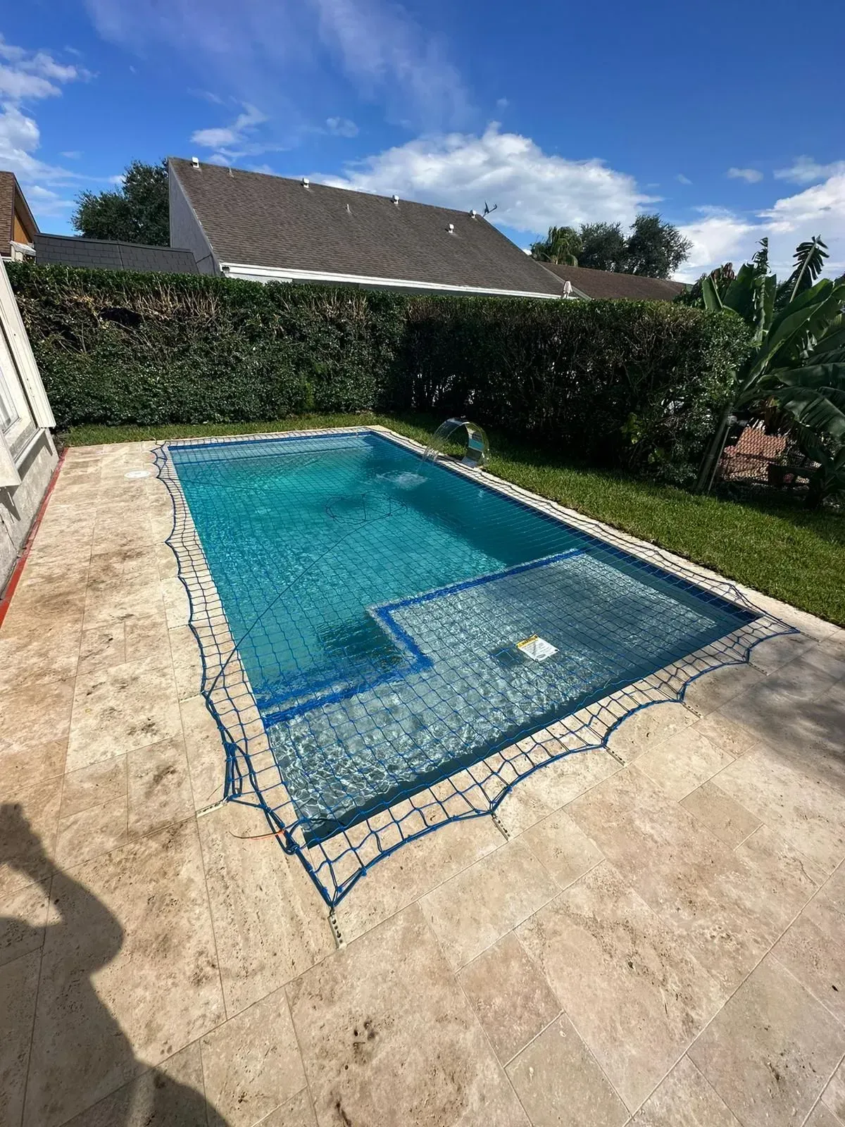 Rectangular swimming pool with blue water and stone patio, surrounded by green shrubs and a clear sky.