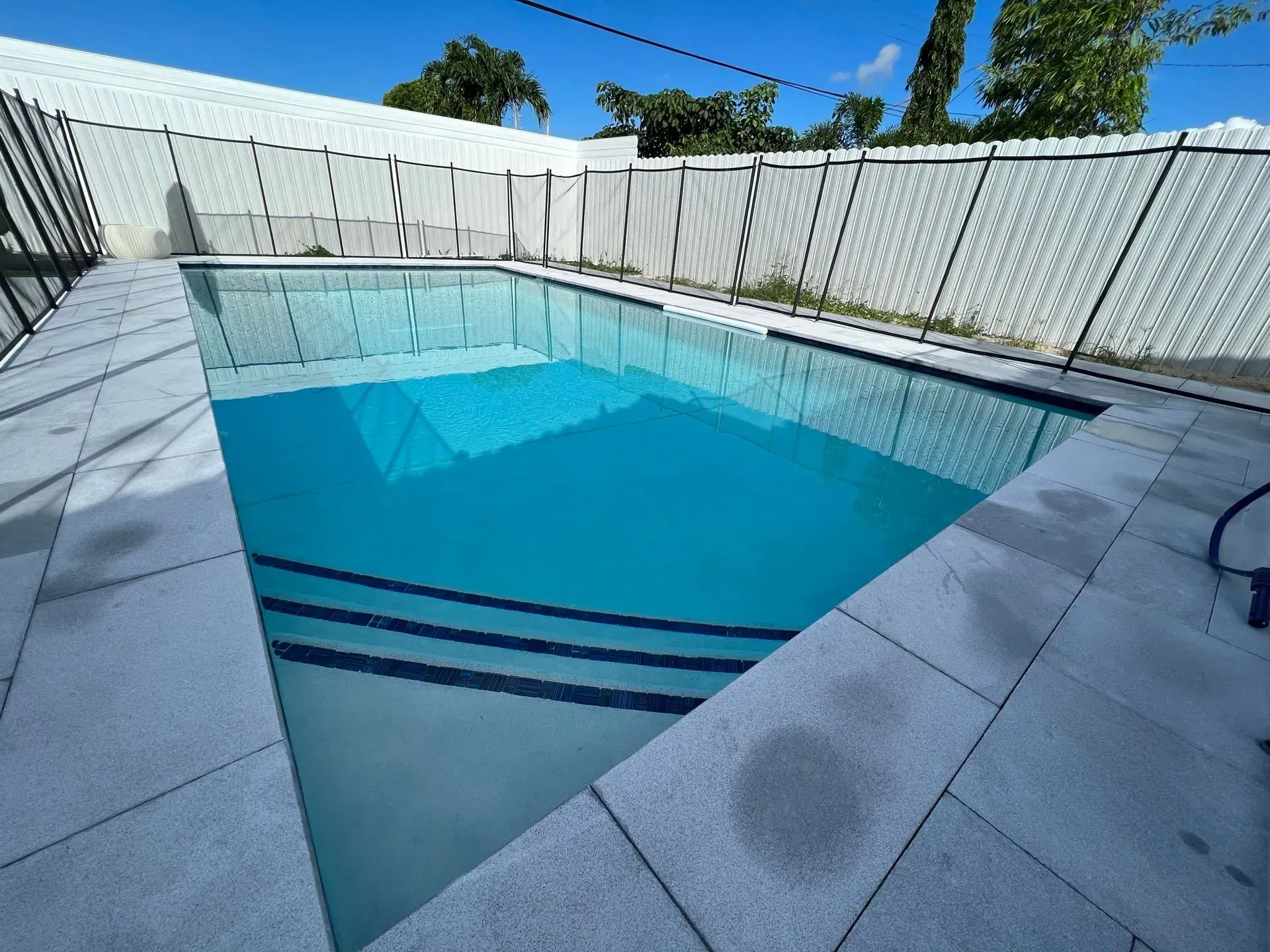 Swimming pool filled with turquoise water, surrounded by light gray tiles and a white fence.