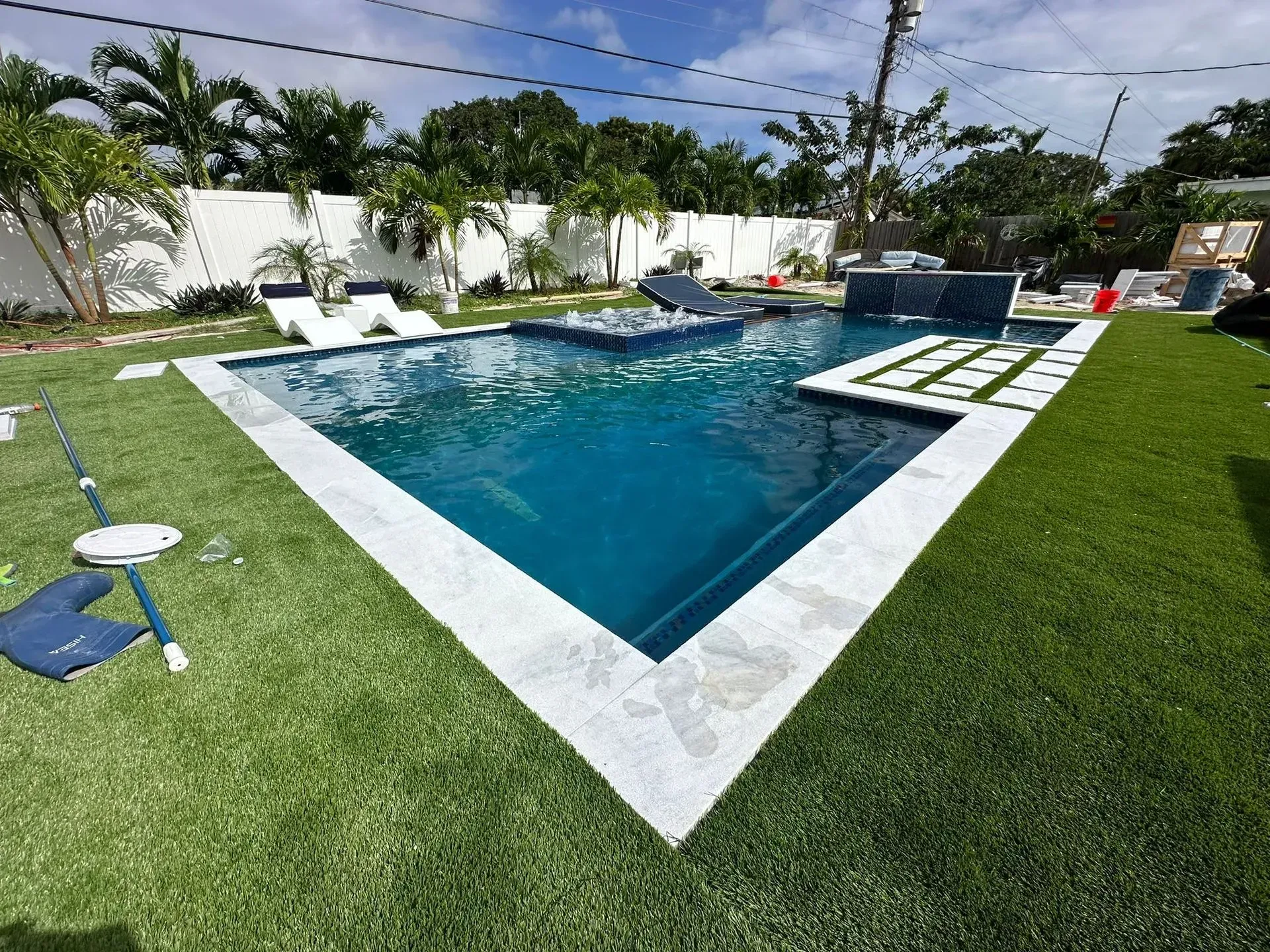 Pool with dark blue water and white tile trim surrounded by green lawn and white fence.