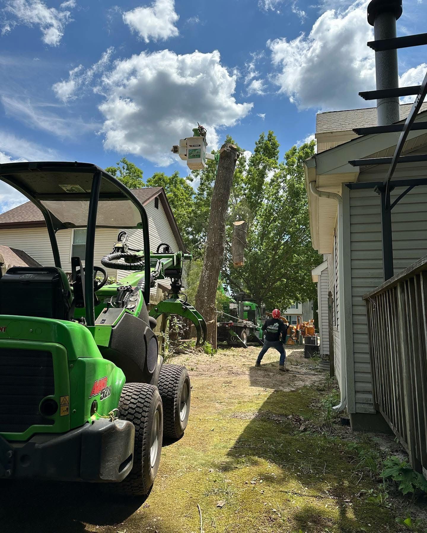 A green tractor is cutting a tree in front of a house.