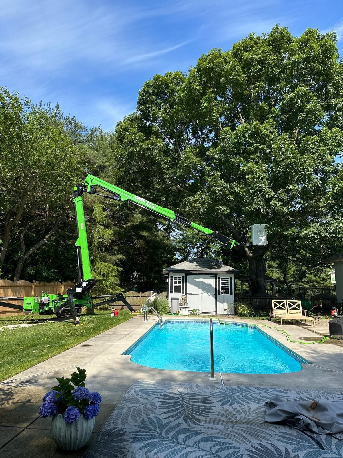 A green crane is cutting a tree next to a swimming pool.