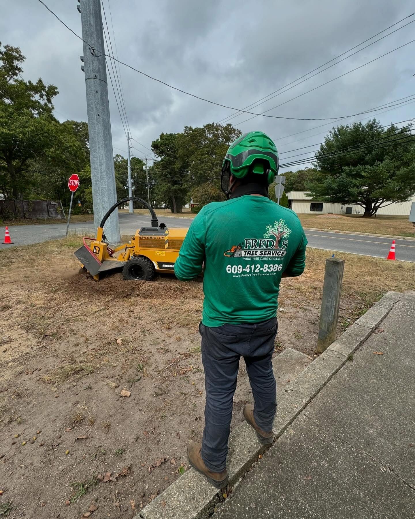 A man in a green shirt is standing next to a tree stump grinder.