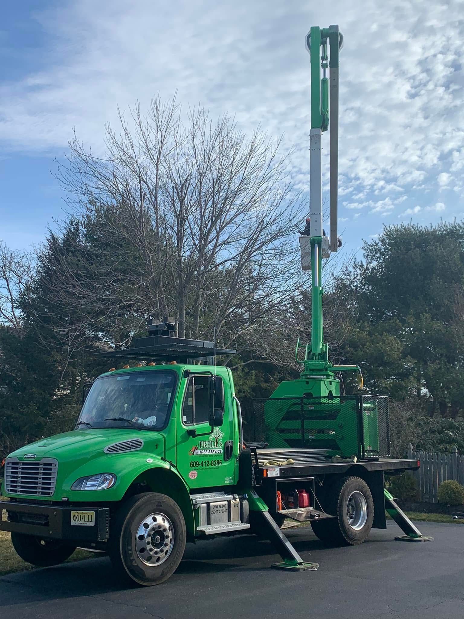 Green truck with a tall crane-like attachment in front of a leafless tree and against a blue sky.