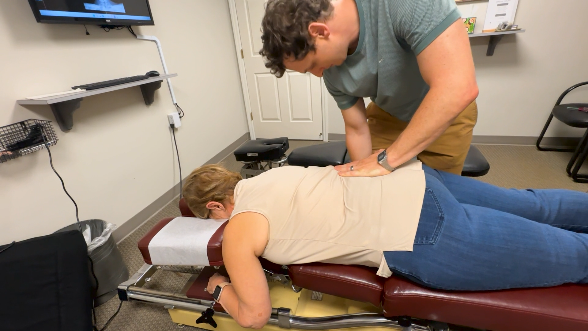 Chiropractor adjusting a patient lying face down on a treatment table in a clinic room.