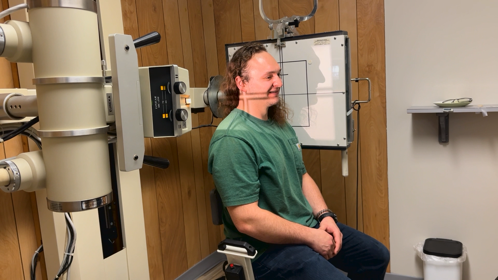 Man seated in a clinical room near medical equipment, wearing a green shirt and headset, smiling in profile.