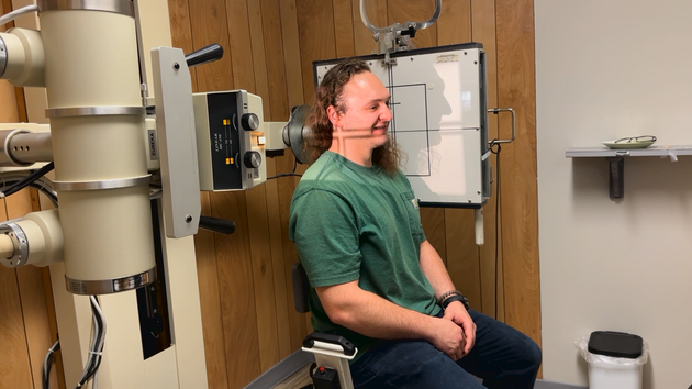 Man seated in a clinical room near medical equipment, wearing a green shirt and headset, smiling in profile.