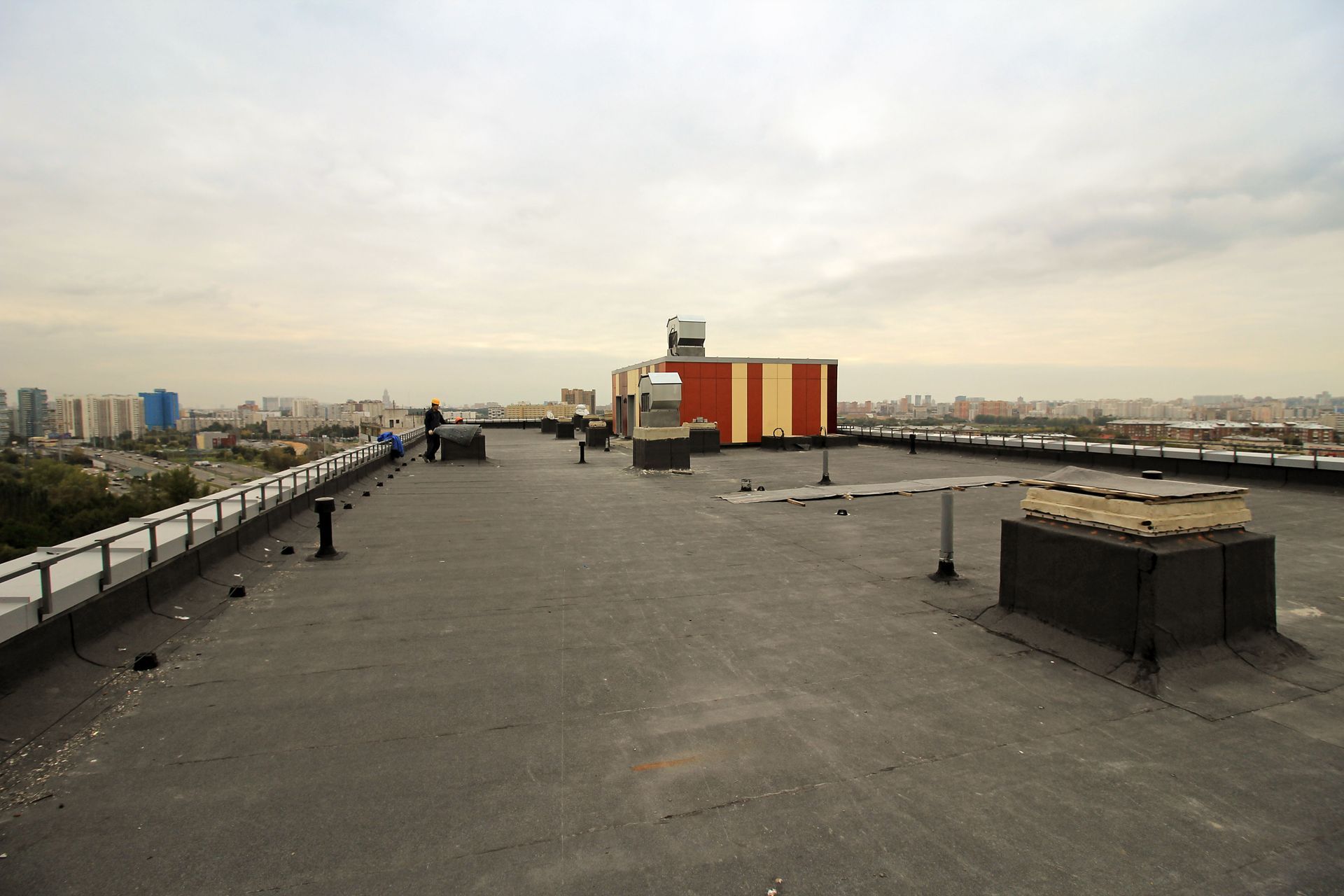 A rooftop with a flat, dark surface, various fixtures, and a distant cityscape under a cloudy sky.