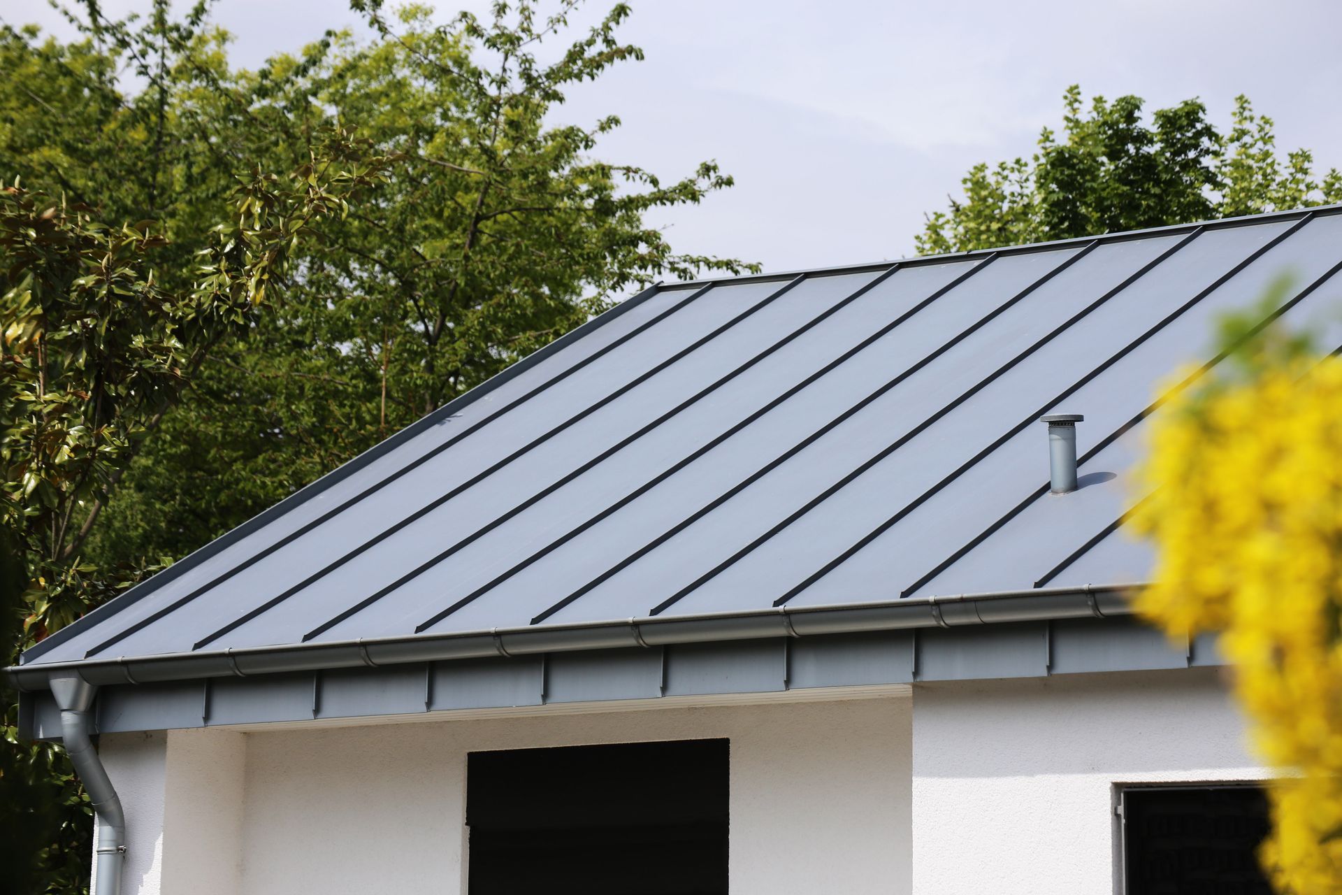 Gray metal roof on a white house with a gutter and chimney, trees in the background.