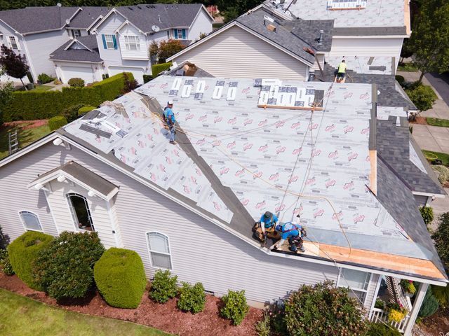 Roofers working on a house roof. Blue uniforms, gray and white roof, sunny day, residential neighborhood.