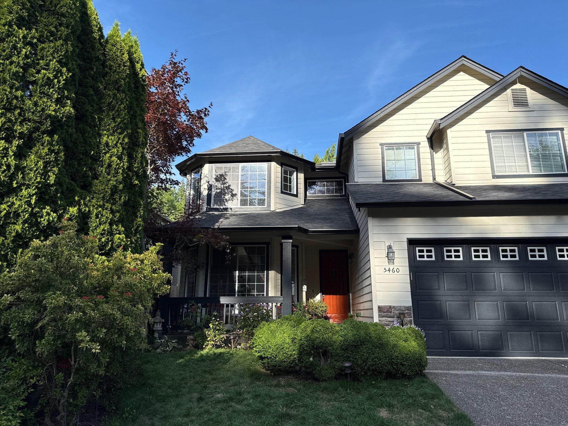 Two-story house with beige siding, a dark garage door, and lush green landscaping under a blue sky.