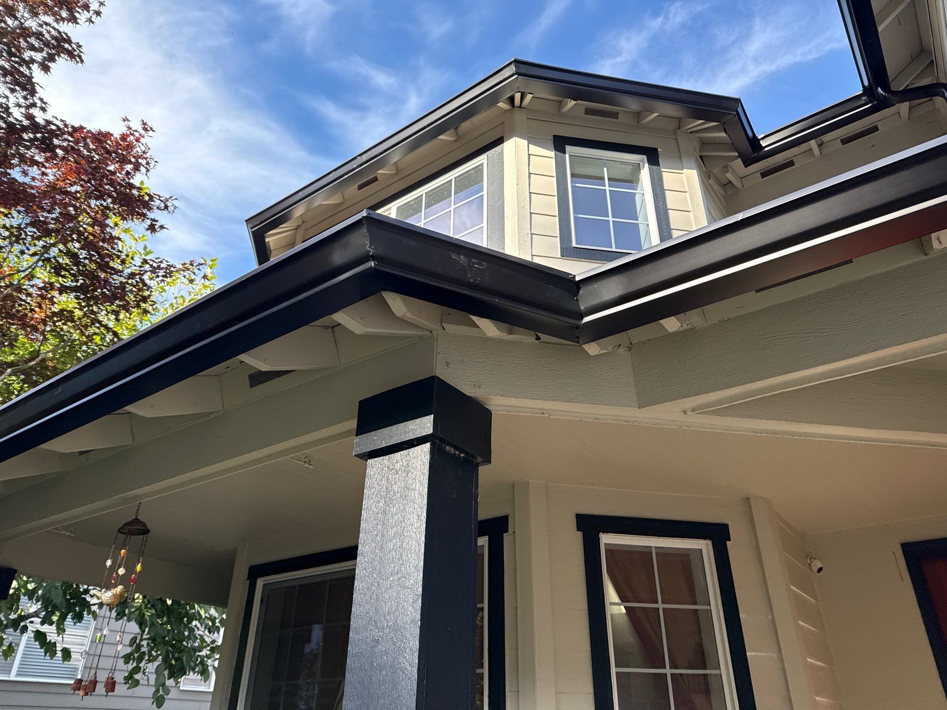 Tan house with black trim, gutters, and pillars. Blue sky with white clouds visible.