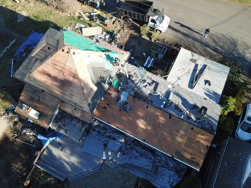 Aerial view of a house undergoing roof replacement; tarps cover parts of the roof; a truck and debris are visible.