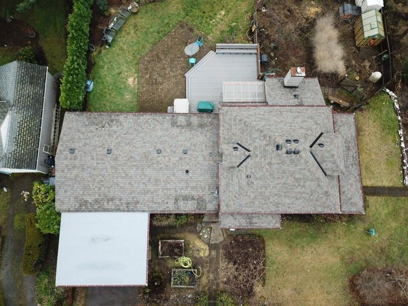 Overhead view of a house with gray roof, attached carport, and backyard with lawn and some greenery.