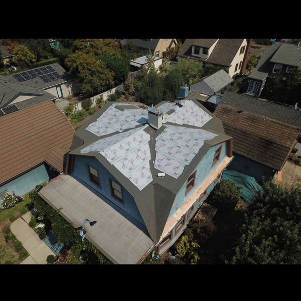 Overhead view of a house with a roof under construction. Gray tarp covers the roof sections; surrounding houses visible.
