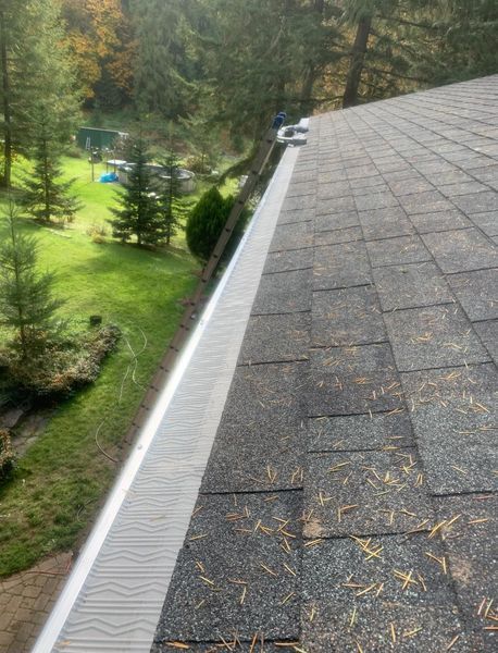 Gutter guard installed along the edge of a roof covered in brown shingles, overlooking a green yard with trees.