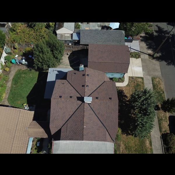 Aerial view of a brown-roofed house with a complex roofline, surrounded by trees and other houses.